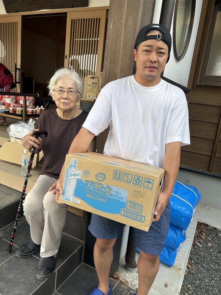 玄関先で、飲料水の箱を運ぶ店主と、受け取られた方